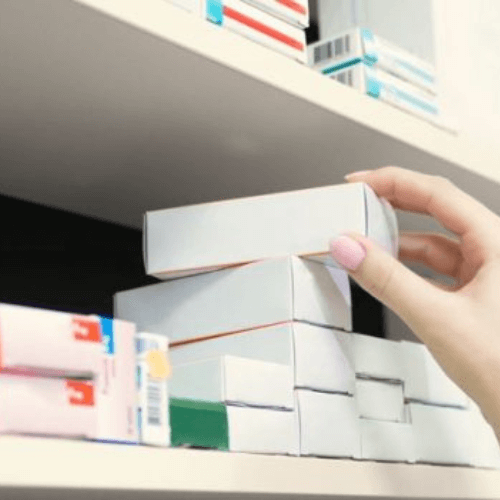 A hand placing a white box on a shelf filled with various medication packages.