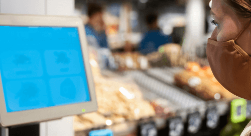 A woman wearing a mask scans bananas at a self-checkout kiosk in a grocery store.